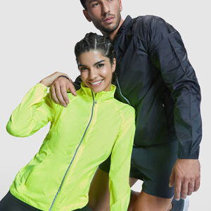 To personer iført træningstøj poserer; kvinden i neongul jakke smiler, og manden i sort jakke står bag hende. Indendørs, lys grå baggrund.