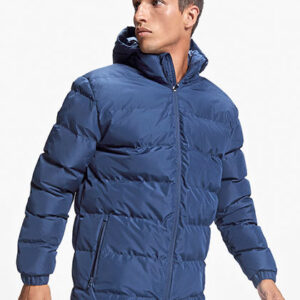 Person iført en blå, vatteret jakke står med hætte oppe i en neutral, lys baggrund. Jakken er quiltet med lynlås foran og to sidelommer.