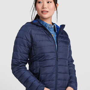 En person bærer en mørkeblå vatteret jakke og en denimskjorte, ser til siden med hænder i lommerne. Baggrunden er ensfarvet lys grå, hvilket fremhæver jakken.