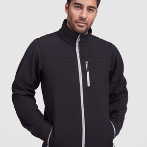 Mand iført sort jakke med hænderne i lommerne, ser direkte fremad. Baggrunden er neutral og lysegrå. Ingen tekst til stede.