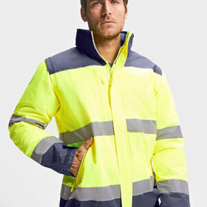 Mand i gul og grå refleksjakke står med hånden i lommen. Han ser beslutsomt fremad i et neutralt, lyst rum.