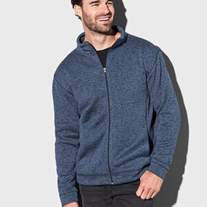 Mand bærer en blå lynlåstrøje, står afslappet med hænderne ved siden, smilende, i et neutralt rum med grå baggrund.