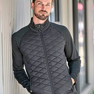 Mand liggende op af en væg iført en mørk quiltet jakke. Han kigger roligt mod kameraet i et udendørs bymiljø.