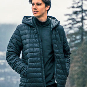 En person iført en quiltet jakke står udendørs, kigger til siden, omgivet af tågede skove og bjerge i baggrunden. Jakken er mørk, og personen har hænderne i lommerne.