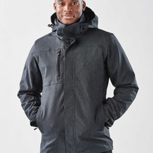 Mand iført mørk jakke med hætte står roligt med hænderne i lommerne på en neutral, lys baggrund.