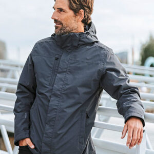 En person iført en mørk jakke står udendørs, kigger til siden med en rolig mine. Baggrund viser en række stole eller bænke og en lidt sløret byscene.