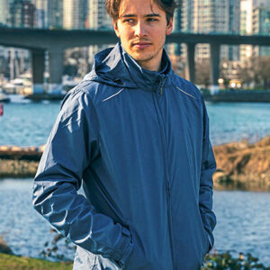 Person i blå jakke står med hænderne i lommerne ved en flod. Baggrunden viser en bro, græs og høje bygninger på afstand i en byscene.