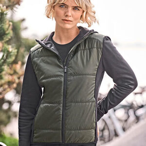 Kvinde iført grøn quiltet vest står afslappet. I baggrunden ses cykler og grønne træer, mens lyset indikerer dagtimerne. Hun har kort blond hår og afslappet kropsholdning.