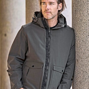 Mand iført jakke står afslappet op ad en søjle i et udendørs miljø. Han kigger til siden og har hænderne i lommerne.