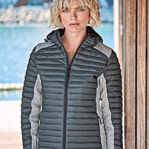 Person med kort, lyst hår står iført en sort og grå dunjakke. Omgivelserne viser en træbeklædt terrasse ved vandet med moderne bygninger i baggrunden.