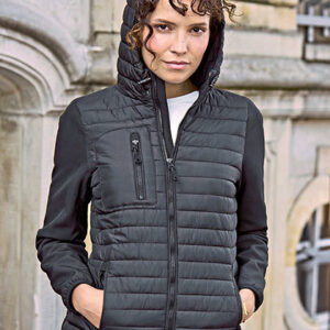 En person iført en sort jakke står afslappet med hænderne i lommerne foran en bygning med buede vinduer og stenarkitektur. Ingen tekst er synlig.