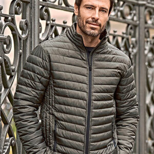 Mand iført en mørkegrøn vatteret jakke står afslappet med hænderne i lommerne foran et udsmykket smedejernshegn i en bymæssig omgivelser.