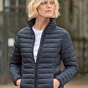 En person med lyst, kort hår bærer en sort, quiltet jakke. Personen kigger til siden med hænderne i lommerne. Baggrunden er sløret, antydende en bymæssig miljø.