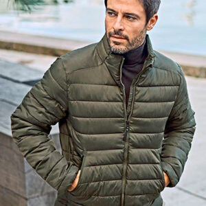 En mand iført en olivengrøn quiltet jakke står med hænderne i lommerne. Han ser til siden i en udendørs omgivelser nær vandet med stenbelægning omkring.