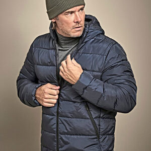 Mand iført blå vatteret jakke med hætte, trækker lynlåsen op. Han bærer en grå strikhue og grå sweater, står mod en beige baggrund.