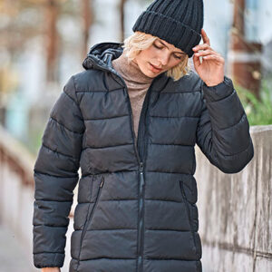 En person i sort dunjakke justerer en mørk strikhue. Personen går i en bymæssig gade med natur og betonvægge omkring sig. Ingen tekst er synlig.