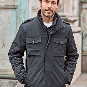 Mand iført mørk jakke står afslappet med hænderne i lommerne foran en rustik murstensvæg.