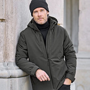 Mand i mørk jakke og hue læner sig mod en bygning i sten i en bymæssig setting. Han ser afslappet ud og kigger fremad.