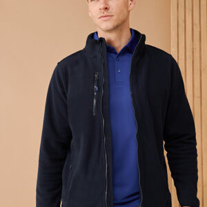 Person iført en sort jakke over en blå trøje, står foran en beige baggrund med lodrette linjer. Jakken er lynet op, og personen kigger fremad.