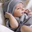 En baby iført en grå hue og dragt ligger afslappet i en hvid, blød dyne, mens den holder let ved sin hue og ser opmærksomt fremad.