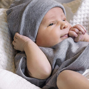 En baby iført en grå hue og dragt ligger afslappet i en hvid, blød dyne, mens den holder let ved sin hue og ser opmærksomt fremad.
