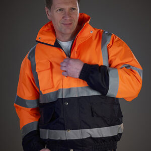 En person iført en orange og sort jakke med reflekterende striber står i en neutral, indendørs belysning. Personen lyner jakken, hvilket antyder klargøring til brug.