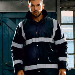 En person iført en mørk jakke med reflekterende striber står i en industriel bygning med blå døre og vinduer i baggrunden.