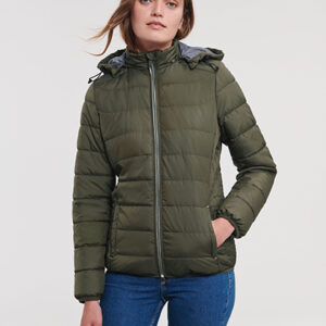 En person iført en olivengrøn, vatteret jakke står oprejst med hænderne i lommerne og ser fremad i et neutralt indendørs miljø. Ingen tekst til stede.