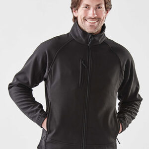 Mand, iført sort jakke med lynlås, smiler; hænder i sidelommer. Han står mod en hvid baggrund, hvilket fremhæver jakken og hans afslappede kropsholdning.