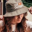 Kvindelig model bærer en lysebrun bøllehat med teksten "Dit Design" i en have. Hun justerer hatten med hånden og har langt, brunt hår.