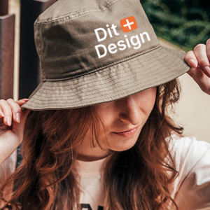 Kvindelig model bærer en lysebrun bøllehat med teksten "Dit Design" i en have. Hun justerer hatten med hånden og har langt, brunt hår.