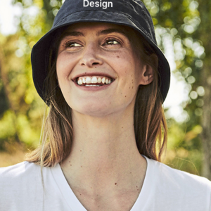 Person smiler, iført sort bøllehat med tekst "Design" og hvid t-shirt, omgivet af grønne træer og sollys i en udendørs park.