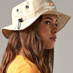 Kvinde iført beige bøllehat ser til siden, med tekst "Dit Design" på hatten. Hun har langt brunt hår og bærer en gul trøje. Baggrunden er grå og ensfarvet.