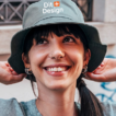En smilende person iført en bøllehat med teksten "Dit Design" justerer hatten med hænderne. Baggrunden viser en mur med grafitti og et vindue, gadeomgivelser.
