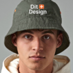 Mand bærer en grøn bøllehat med teksten "Dit Design" foran, iført en beige hættetrøje, stående i et neutralt, gråt rum.