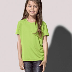 En ung pige står smilende iført en lysegrøn T-shirt og mørke bukser i en neutral indendørs setting med en skygge på væggen bag hende.